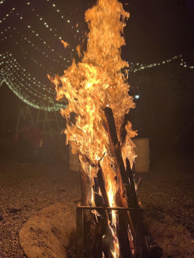 Una fogata con leña ardiendo en un contenedor metálico, rodeada de grava y luces colgantes al fondo.
