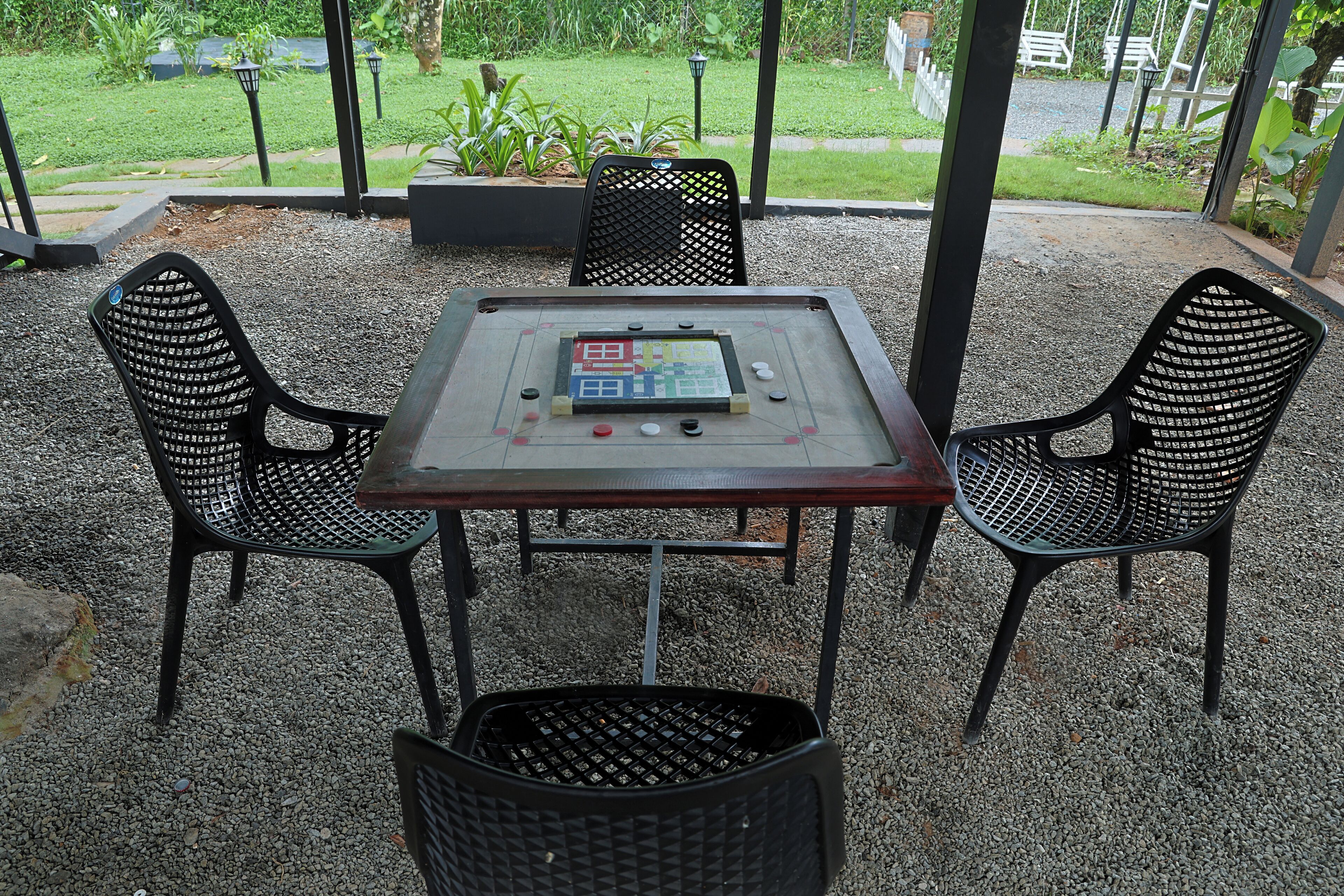 A table with a board game, surrounded by four chairs, set on a gravel surface with a garden view.