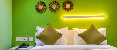 Habitación de hotel con pared verde, una cama con ropa de cama blanca y dos almohadas, una mesita de noche con teléfono y una lámpara fijada a la pared.