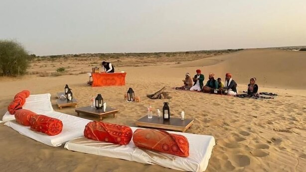 Paisaje desértico con un grupo de personas sentadas sobre una manta, una mesa con comida y bebidas, y una tienda de campaña con alguien adentro.