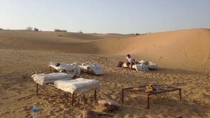 Un campamento en el desierto con tiendas de campaña, una mesa con comida y una persona sentada sobre una duna de arena.