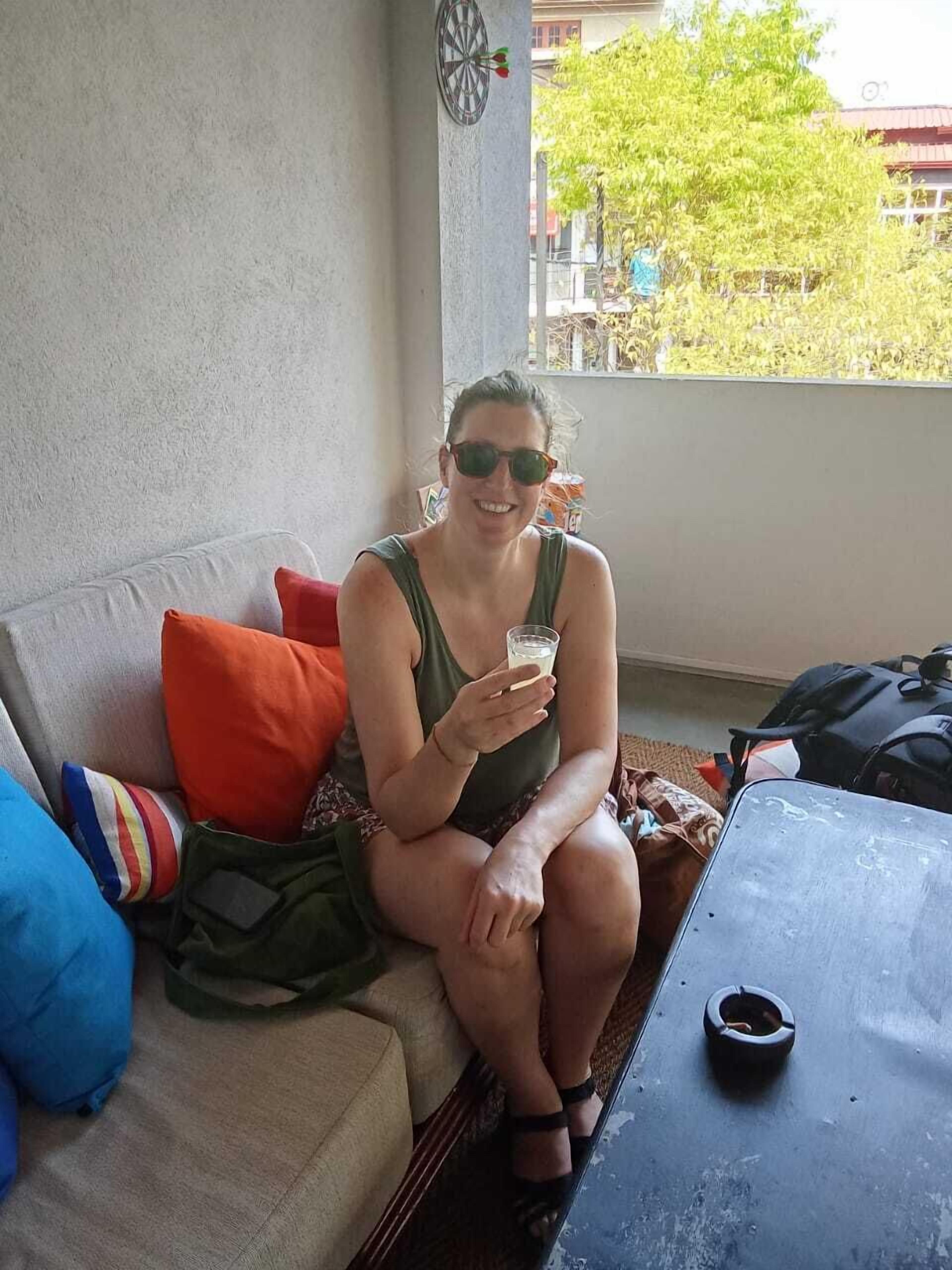 A person sitting on a sofa with a backpack and a glass of drink, a dartboard on the wall, and a view of buildings and trees outside.