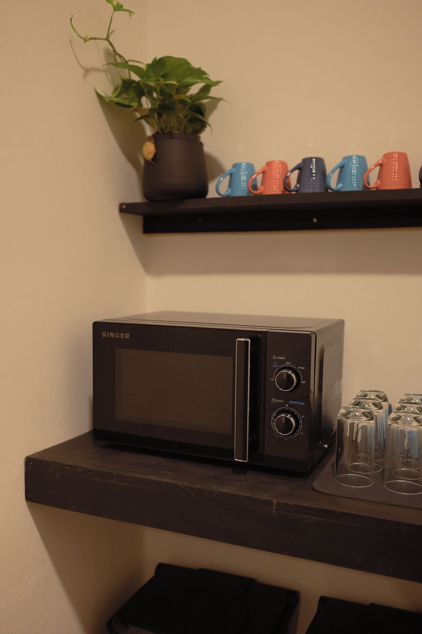 A black microwave on a dark countertop, a shelf with colorful mugs, and a potted plant.