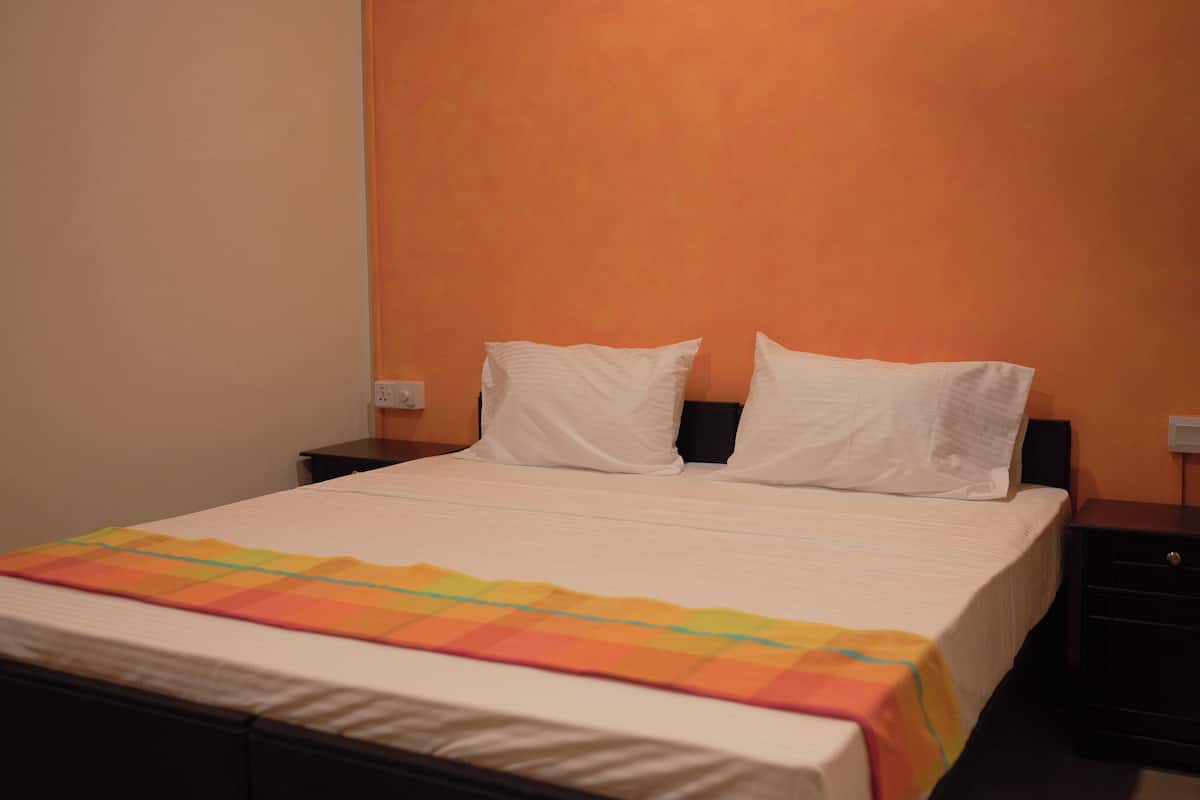 Ein Bett mit weißer Matratze und Kissen, eine bunte Decke und ein schwarzer Nachttisch mit Lampe.