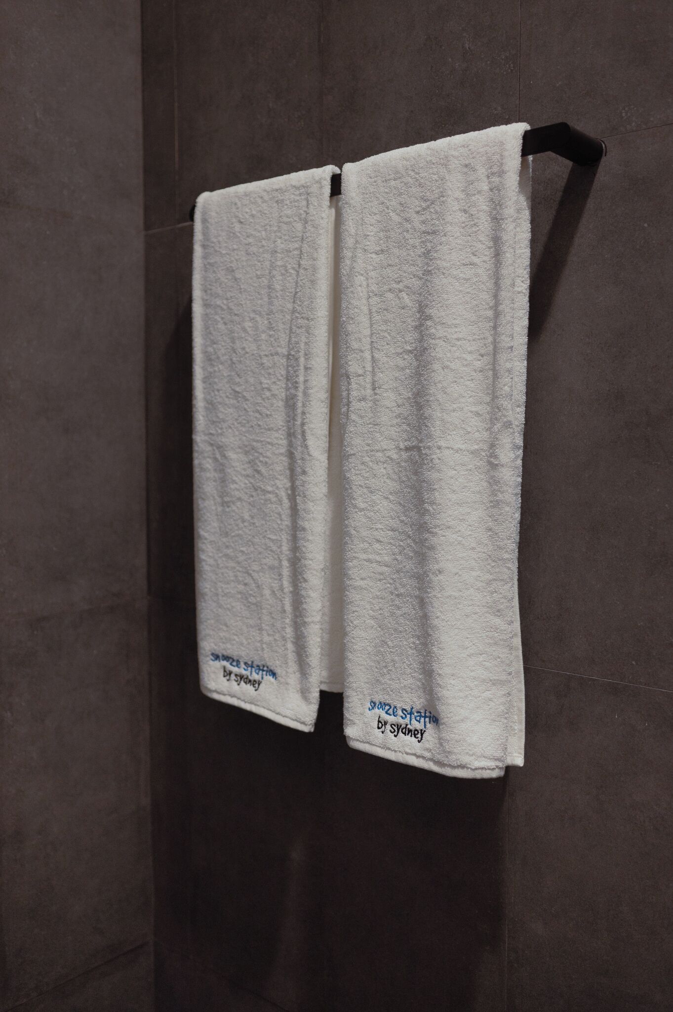 Due asciugamani bianchi appesi a un portasciugamani nero in un bagno con piastrelle scure.