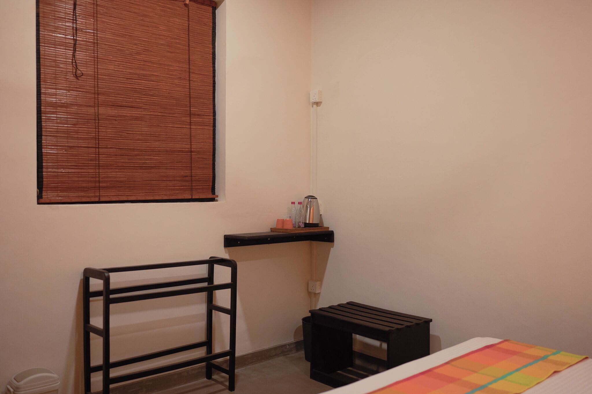 A small, simple room with a bed, a bench, a shelf with toiletries, and a window with blinds.