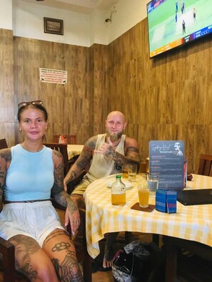 A room with wooden walls and a television mounted on the wall displaying a sports event. There is a table with a yellow checkered tablecloth, a menu stand, and a bottle of beer. - Snooze Station by Sydney Galle (Galle)