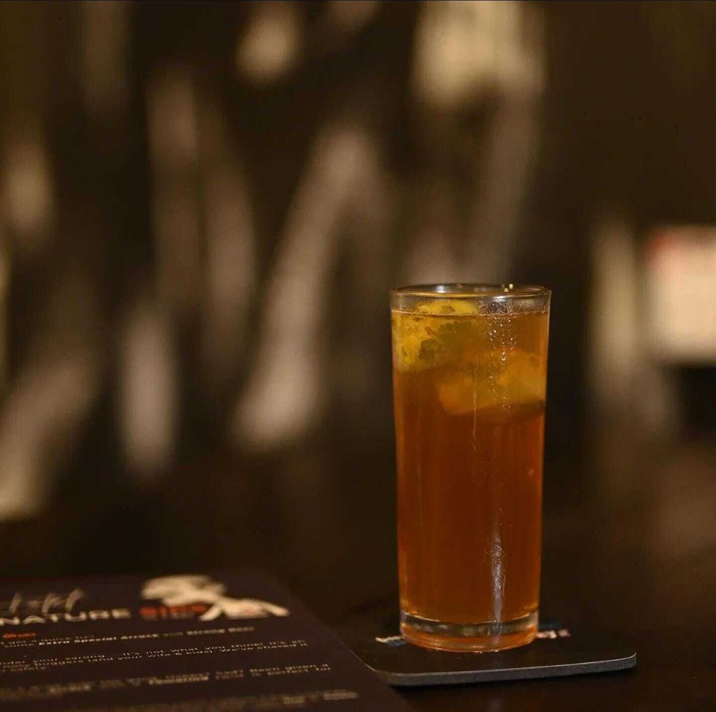 A glass of amber-colored beverage on a coaster with a blurred background.