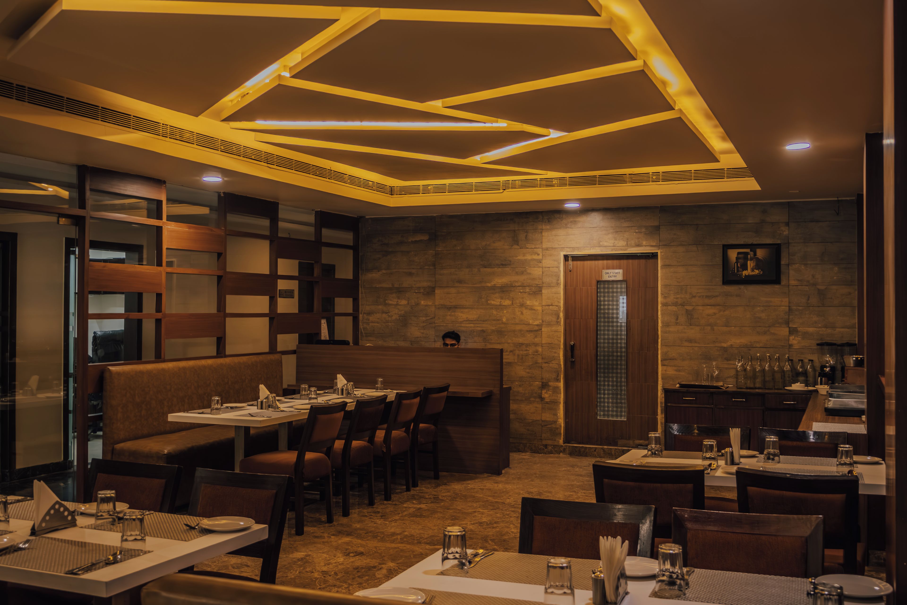 Ein Restaurant mit modernem Interieur, das sich durch eine hohe Decke mit geometrischer Beleuchtung, Holzpaneele und unterschiedliche Sitzmöglichkeiten, darunter Sitzecken und Tische, auszeichnet.