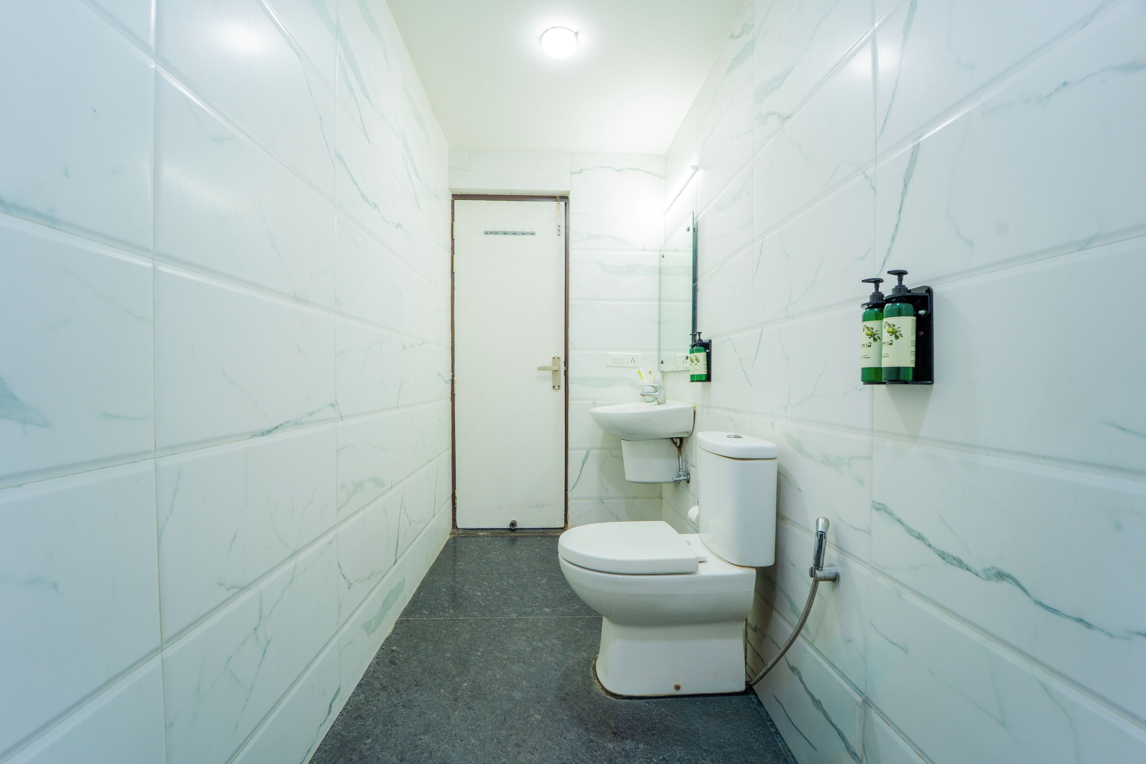 A narrow bathroom with white marble walls, a white toilet, a sink, and two green bottles on a wall-mounted dispenser.