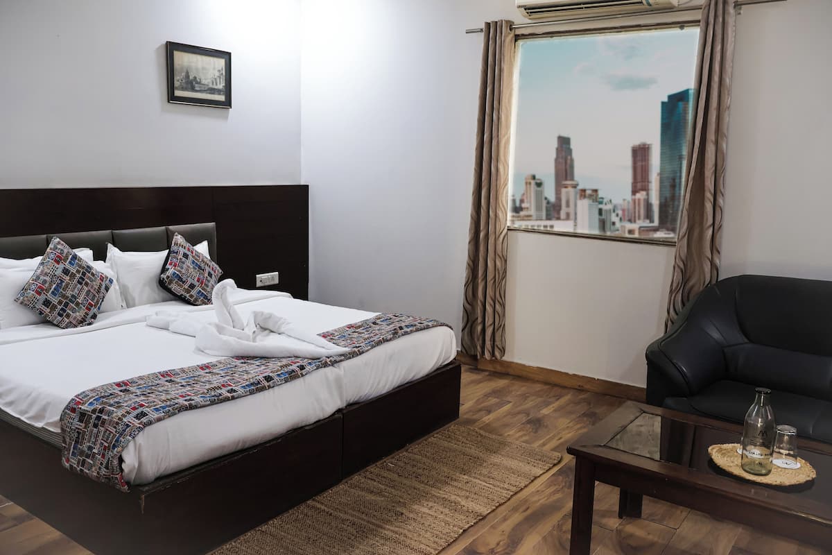 A hotel room with a bed, a black leather sofa, a glass coffee table, a view of a cityscape through a window, and a framed picture on the wall.