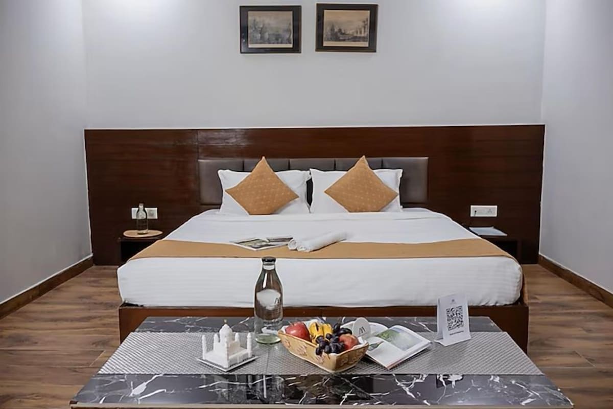 A hotel room with a large bed, two nightstands, a marble table with a fruit basket, a bottle, and a QR code sign.