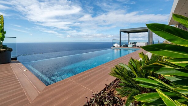 A modern infinity pool with a clear view of the ocean, a covered seating area, and lush greenery.