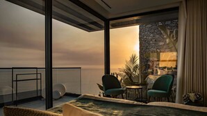 A modern bedroom with a large bed, a seating area with two chairs, a small table, and a view of the ocean through large windows.