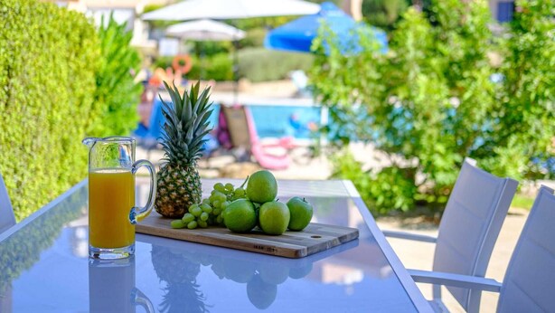 A table with a glass of orange juice, a pineapple, and a wooden board with green apples and grapes.