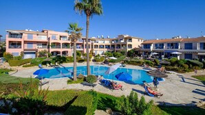 A resort with a central swimming pool surrounded by lounge chairs and umbrellas, multiple-story buildings with balconies, and lush landscaping.