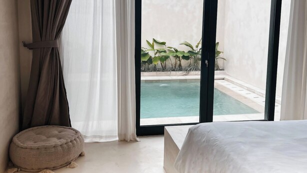 A bedroom with a bed, a window with curtains, and a view of a pool. - Lilla Villa by Betterplace (Canggu)