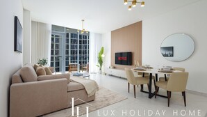 A modern living room with a sofa, dining table, and chairs, a TV, and a large window. - LUX | 2BR Lagoon View – District One, 10–15 min Burj Khalifa (Dubai)