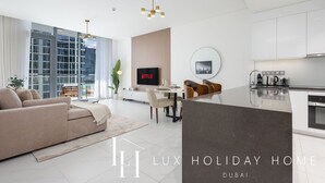 A modern, well-lit living room with a sofa, dining area, and kitchen. A flat-screen TV is mounted on the wall, and there is a large window providing natural light. - LUX | 2BR Lagoon View – District One, 10–15 min Burj Khalifa (Dubai)