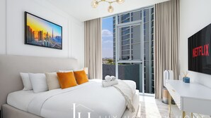 A modern hotel room with a large bed, a cityscape painting, a balcony with curtains, a desk with a Netflix sign, and a view of buildings. - LUX | 2BR Lagoon View – District One, 10–15 min Burj Khalifa (Dubai)