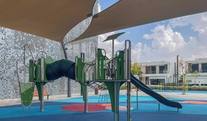 A playground with climbing structures and slides, set against a backdrop of modern buildings. - LUX | 2BR Lagoon View – District One, 10–15 min Burj Khalifa (Dubai)
