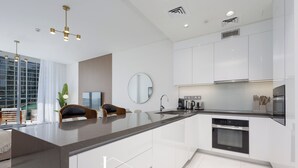 A modern kitchen with white cabinetry, a central island, and a large window providing natural light. - LUX | 2BR Lagoon View – District One, 10–15 min Burj Khalifa (Dubai)