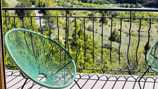 A balcony with two green chairs, offering a view of a valley with mountains and a river.