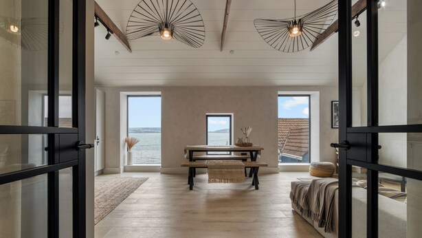 A modern interior with a wooden floor, a central table with a bench, and large windows offering a view of the sea. - Coastal Penthouse, Panoramic Sea View Apartment (The Mumbles)