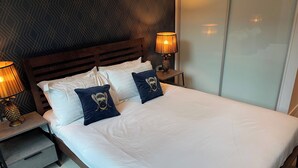 Una cama bien hecha con sábanas blancas y dos almohadas, una con diseño de abeja. Hay dos mesitas de noche con lámparas, una de ellas con un adorno de piña. La habitación tiene un papel tapiz de estampado geométrico y un espejo grande en la pared.