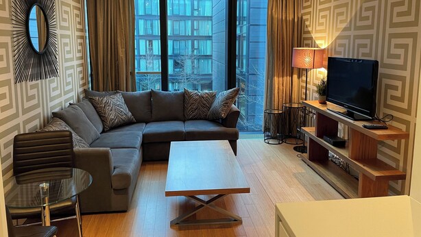 Una sala moderna con un sofá modular, una mesa de centro de vidrio, un televisor sobre un mueble de madera y vistas de un edificio a través de grandes ventanales.