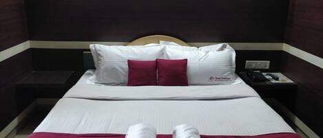 A hotel room with a bed, two pillows, a red blanket, two rolled towels, a nightstand with a phone, and wall-mounted lights.