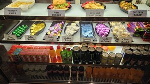 Eine Vitrine mit verschiedenen Speisen, darunter Pommes, ein Gemüseburger, ein Sandwich mit verschiedenen Gemüsesorten und ein Proteinsalat. Darunter befinden sich verschiedene Schokoladensorten und eine Auswahl an Erfrischungsgetränken.