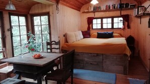 A cozy cabin room with a wooden interior, a bed with pillows, a table with a fruit bowl, and a window with curtains. - Circus wagon star tent with sauna. Very spacious with pavilion. (Windeck)