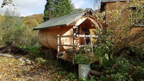 A rustic wooden cabin with a sloped roof, surrounded by dense foliage and a large cylindrical tank. - Circus wagon star tent with sauna. Very spacious with pavilion. (Windeck)
