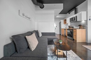 Una sala moderna con un sofá seccional gris, una mesita de centro de madera y una cocina con electrodomésticos de acero inoxidable.
