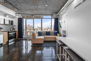 A modern apartment with a kitchen, a living area with a sofa and a dining table, large windows with a city view, and a concrete ceiling. - Calgary Tower Views | Bright Studio | AC (Calgary)