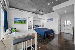 A modern room with a bed, a desk, and a chair. There is a window with curtains, a painting on the wall, and a round mirror. - Calgary Tower Views | Bright Studio | AC (Calgary)