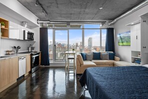 A modern hotel room with a kitchen, a sofa, a bed, and a city view.