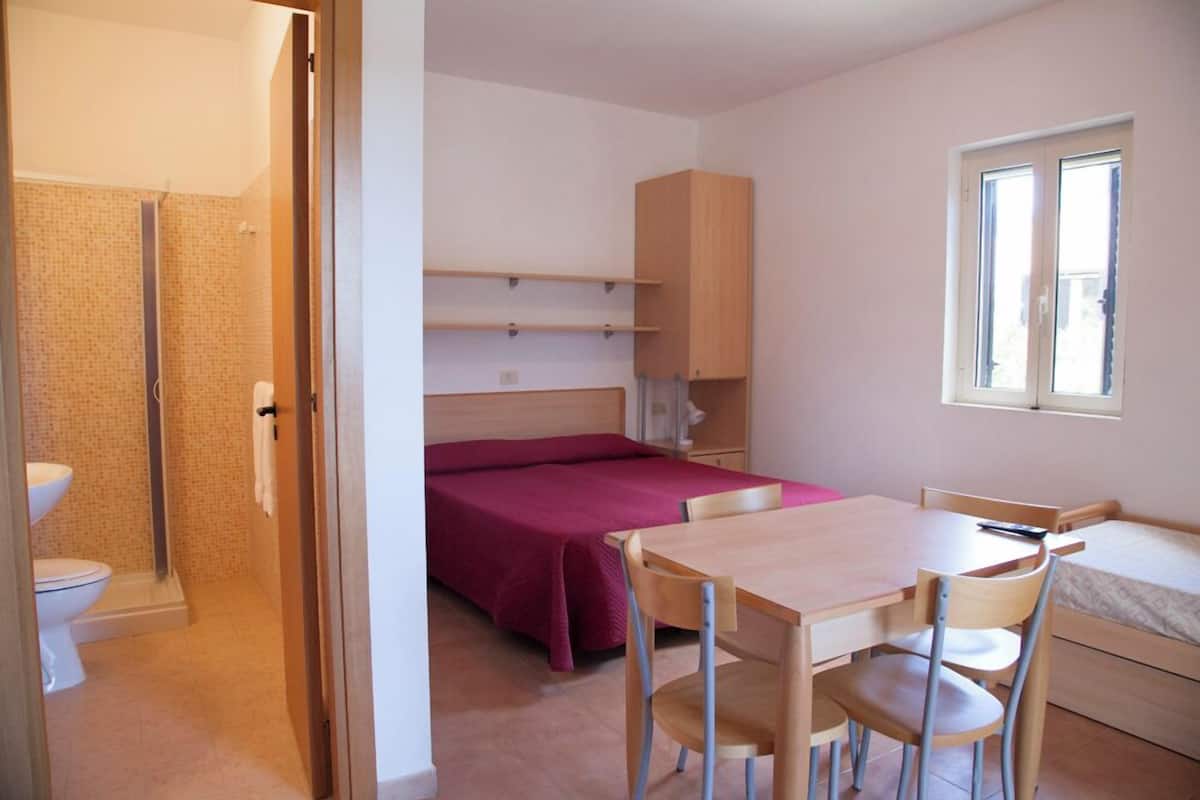 A small, well-lit room with a bed, a desk, a chair, a window, and a shower area.