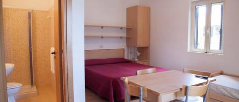 A small, well-lit room with a bed, a desk, a chair, a window, and a shower area.