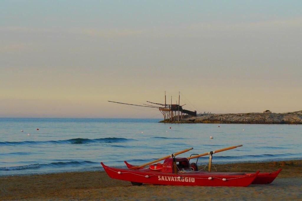 Un bote rojo llamado SALVATAGGIO en una playa arenosa con aguas tranquilas y un muelle distante.