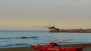 Un bote rojo llamado SALVATAGGIO en una playa arenosa con aguas tranquilas y un muelle distante.