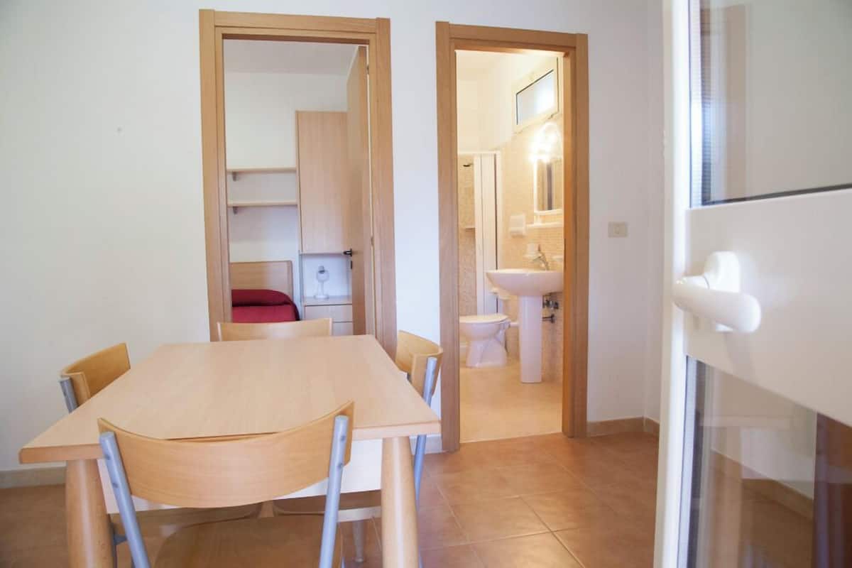 A compact room with a wooden table and chairs, a small kitchenette with a sink, and a toilet visible through an open door.