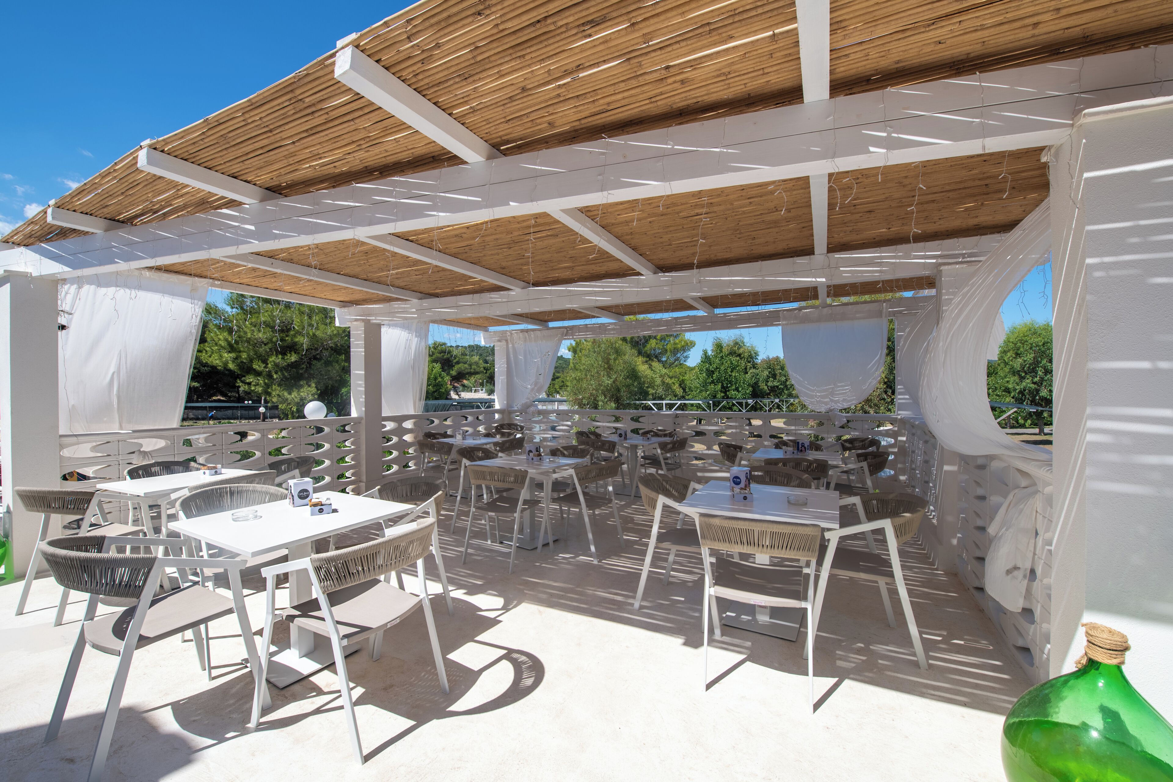 An outdoor patio area with white tables and chairs, covered by a wooden roof structure.