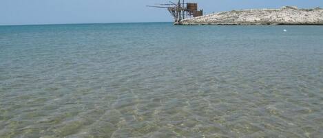 Una zona costera despejada con una estructura que se adentra en el agua, posiblemente un muelle o un embarcadero, y una playa arenosa al fondo.