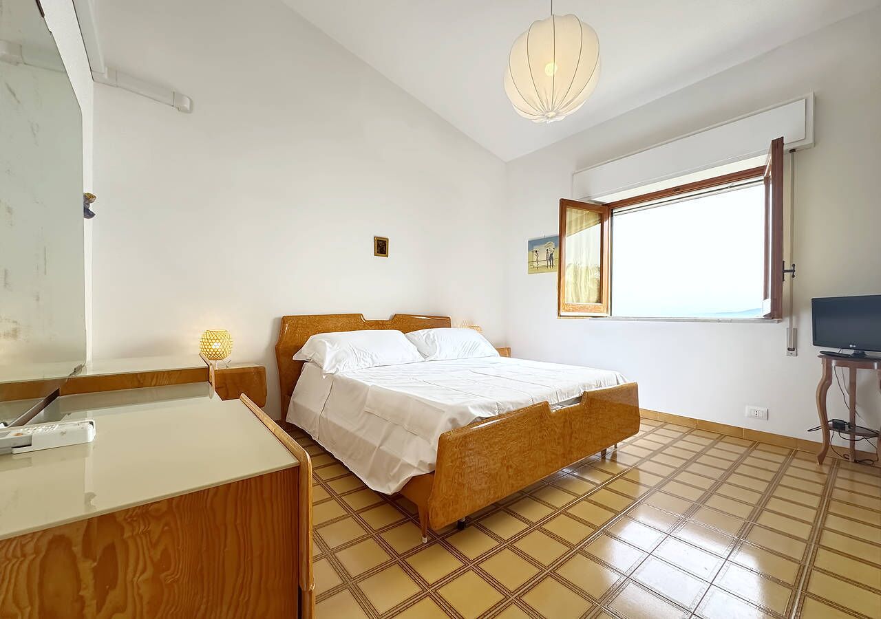 A bedroom with a bed, a desk, a chair, a lamp, a television, and a window.