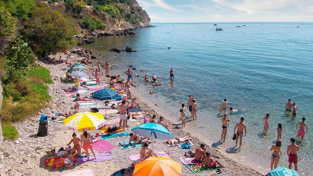 Una spiaggia con persone che prendono il sole e nuotano, ombrelloni e barche in acqua.