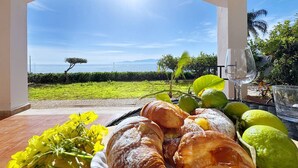 Area pranzo all'aperto con vista sul mare, con un tavolo imbandito di pane, frutta e un bicchiere d'acqua.