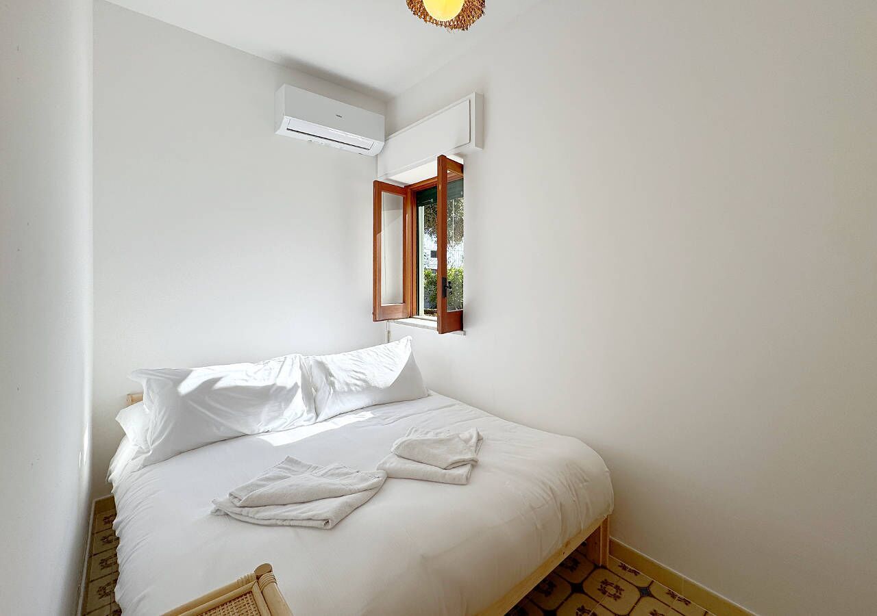 Una cama bien hecha con sábanas blancas y una toalla doblada, una ventana con marco de madera y un aire acondicionado de pared.