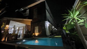 A nighttime view of a hotel with a swimming pool, a well-lit entrance, and a modern architectural design. - Batukaru Garden 5 Bali Sevilla Residence (Tumbak Bayuh)